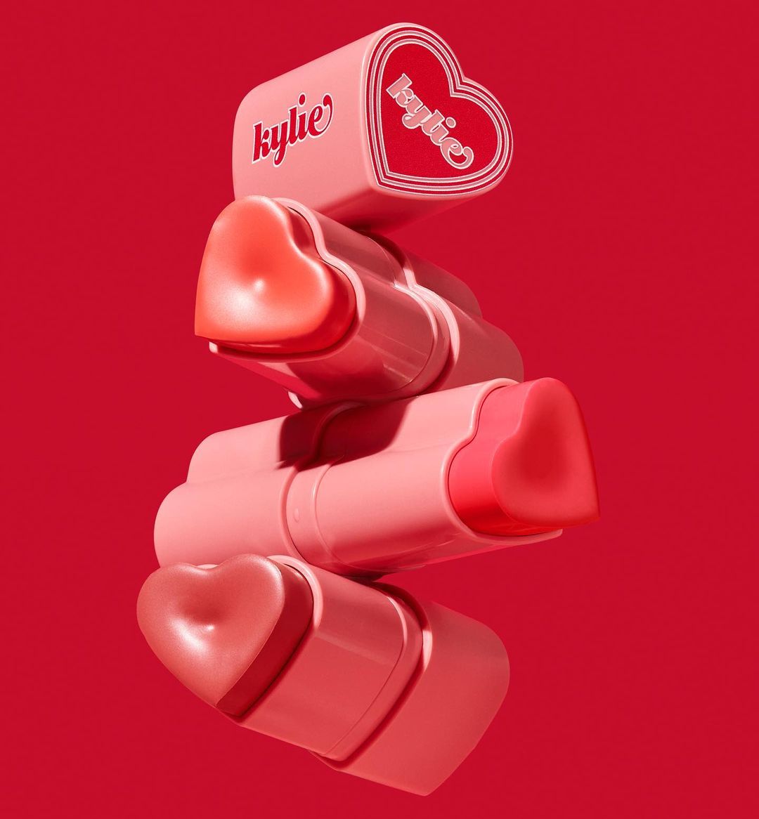Stacked Kylie Cosmetics lip balms with heart-shaped tops against a vibrant red backdrop, product photography for beauty and fashion.