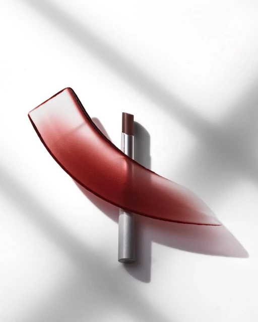 Close-up of a brown lipstick tube with a curved clear and red plastic accent piece on a white surface with plant shadows.