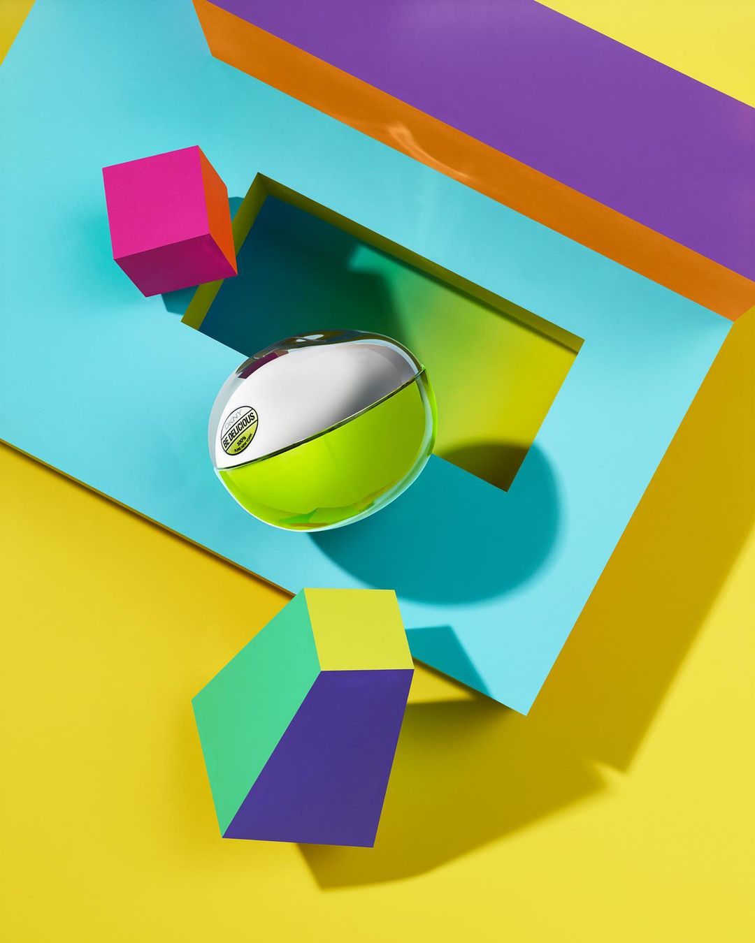 Still life of a lime green and silver spherical bottle amongst colorful 3D geometric shapes on a vibrant yellow backdrop. 