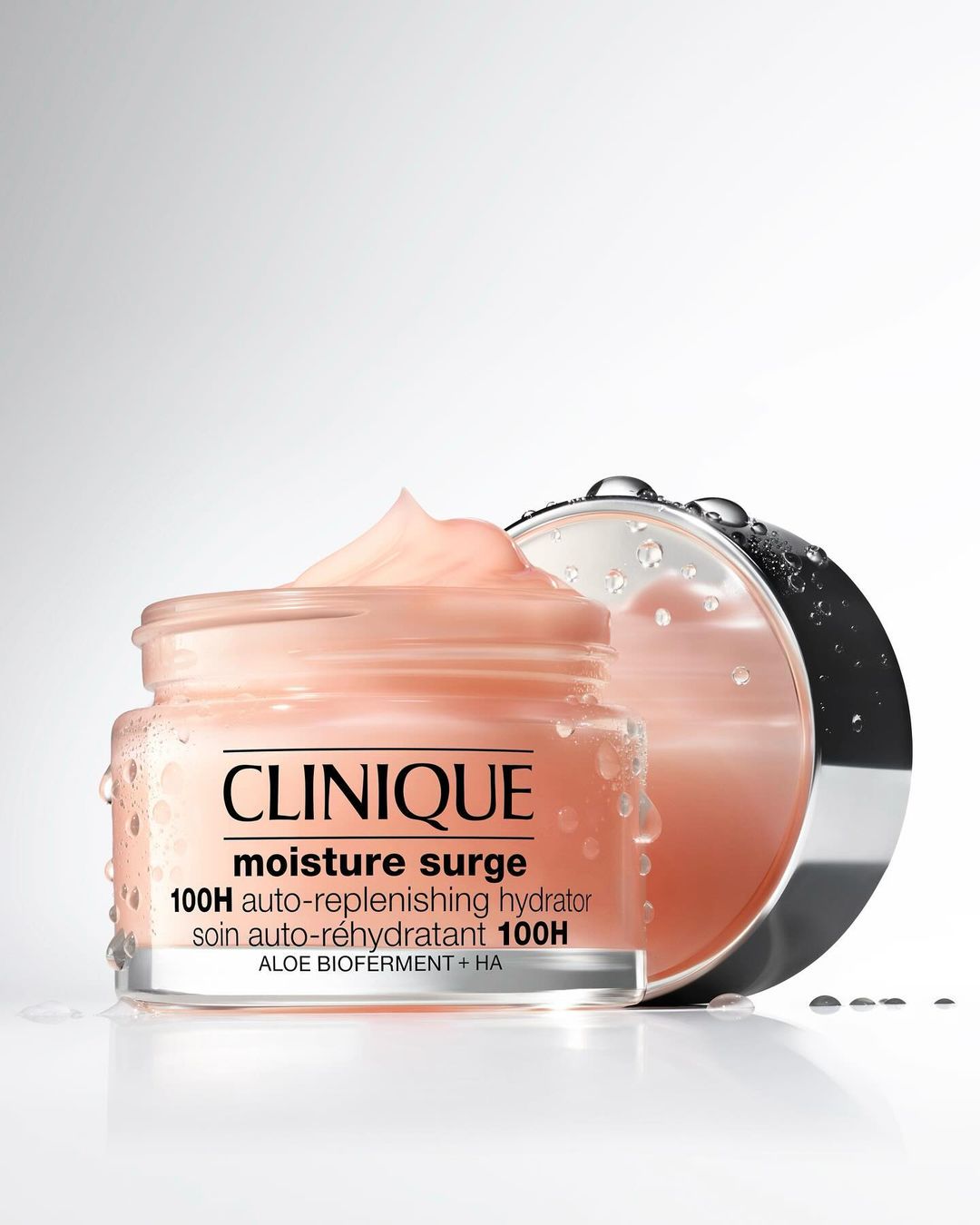Clinique Moisture Surge 100H auto-replenishing hydrator in a clear jar with lid, set against a graduated white background with water droplets.
