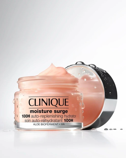 Clinique Moisture Surge 100H auto-replenishing hydrator in a clear jar with lid, set against a graduated white background with water droplets.