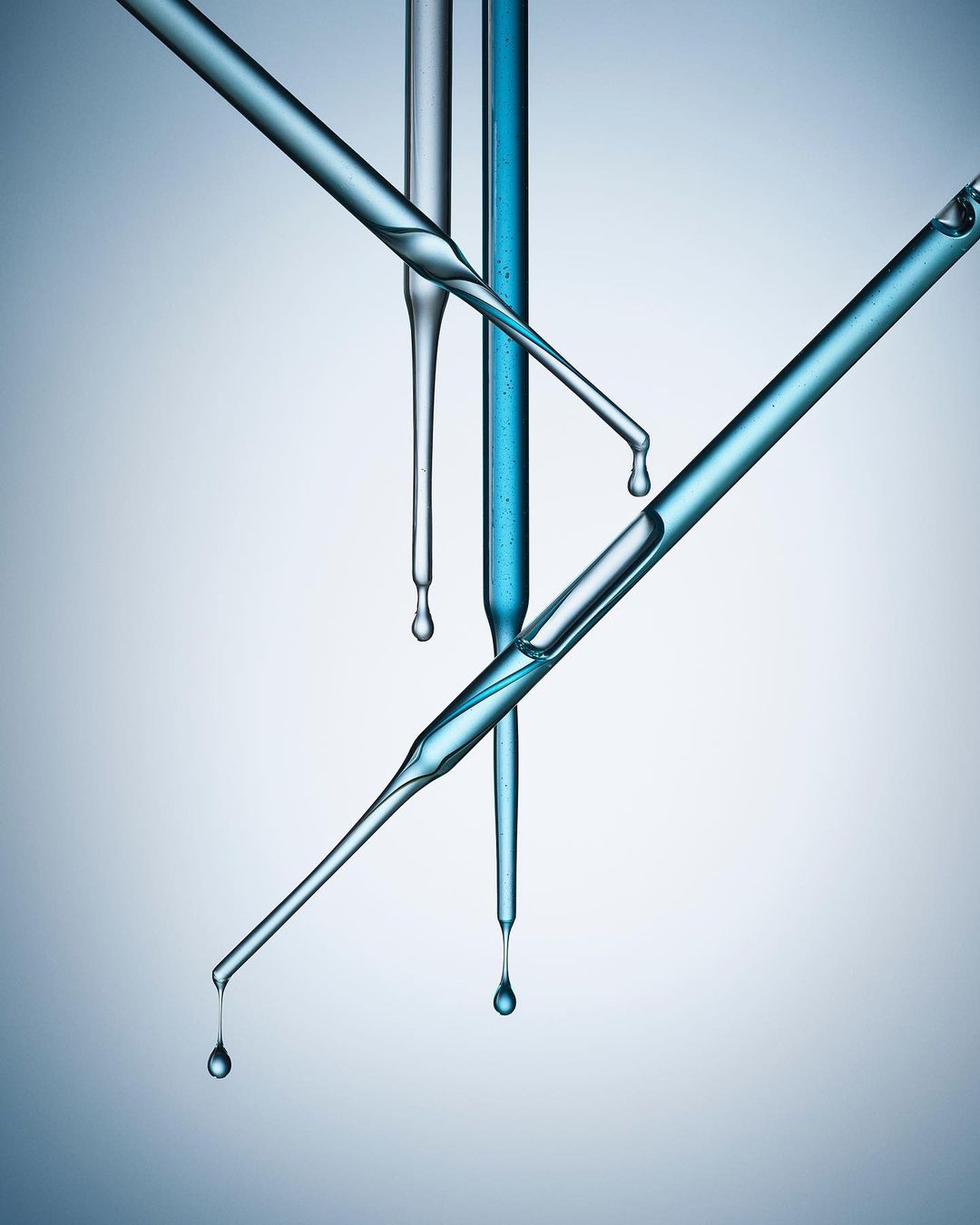 Close-up studio shot of laboratory glass pipettes with blue liquid droplets, set against a blue gradient background.