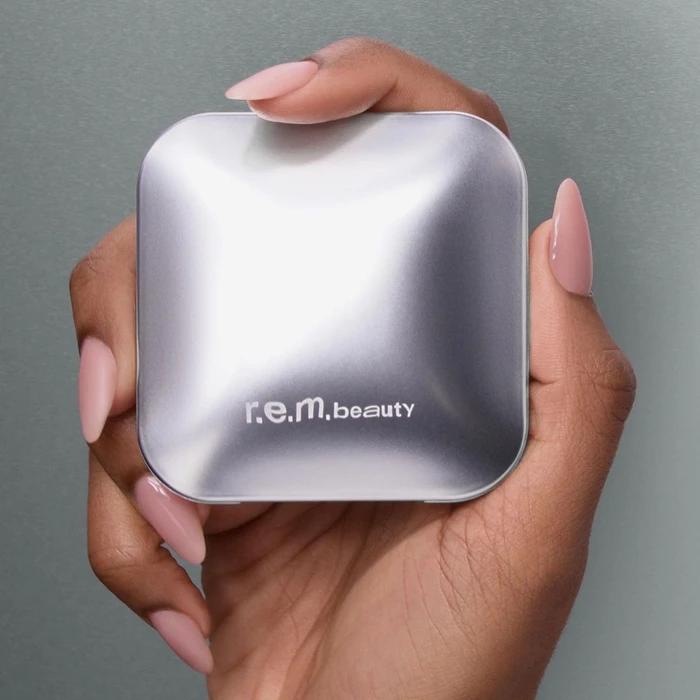 Close-up of hands holding a silver square cosmetic compact with light pink almond-shaped nails against a grey backdrop