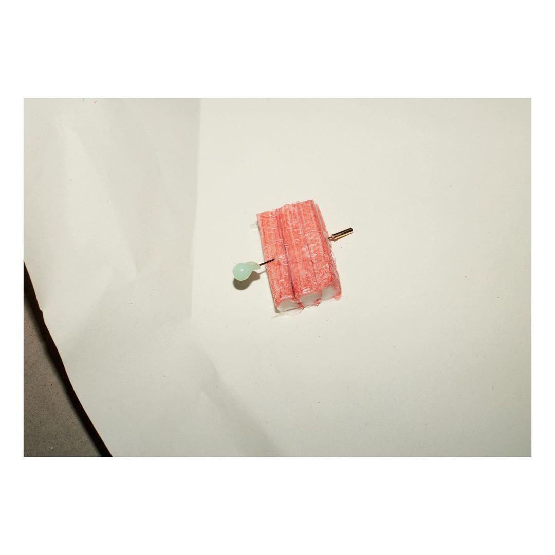 Crab sticks pierced by a straight pin with a green bead head, arranged on a white surface with soft lighting and subtle shadow detail.