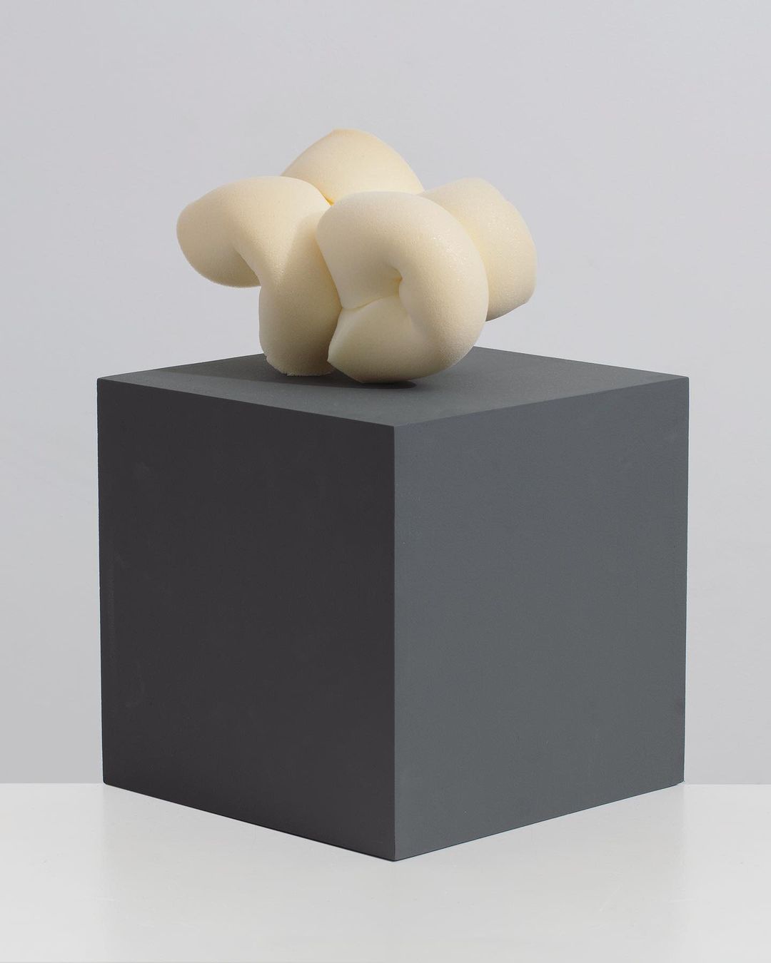 Abstract art piece featuring a pale yellow, organic form sculpture on a dark grey cubic pedestal against a pale grey background.