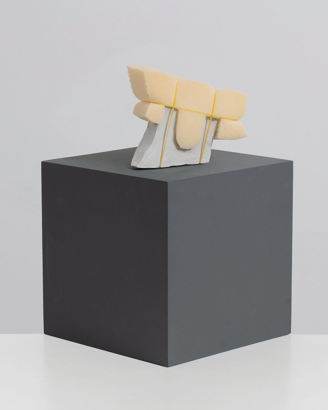 Abstract sculpture features tan foam blocks secured to a light grey concrete base with yellow rubber bands, displayed on dark grey block.