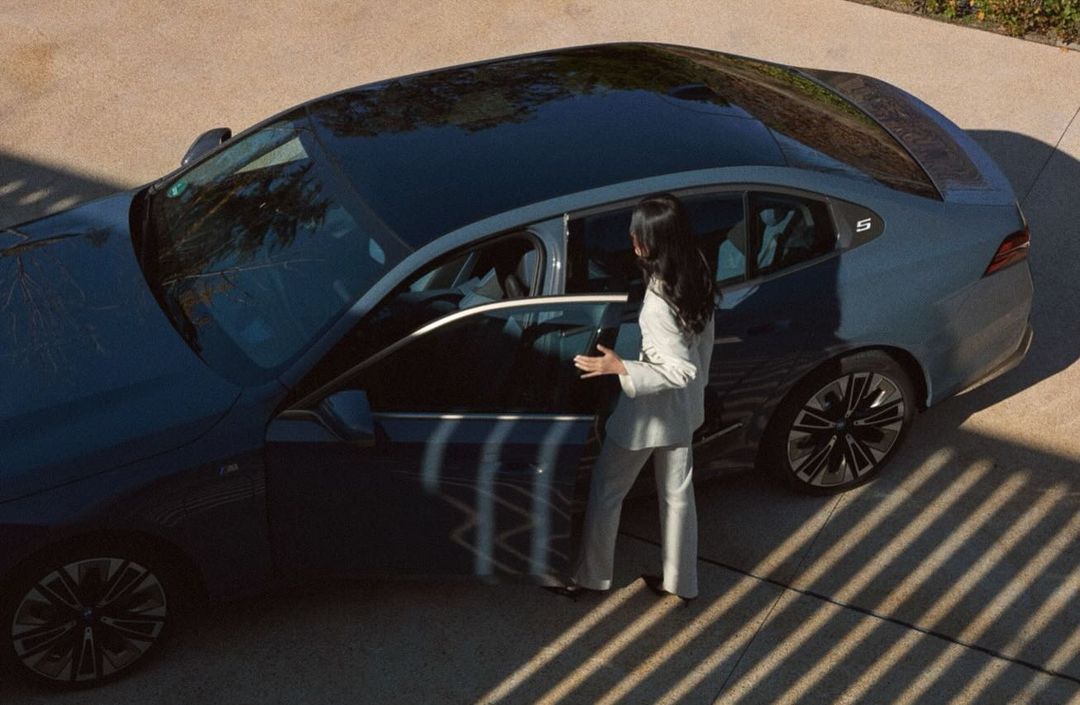 Woman in white suit opening the door of a dark blue car on a paved surface with light and shadow creating a pattern.
