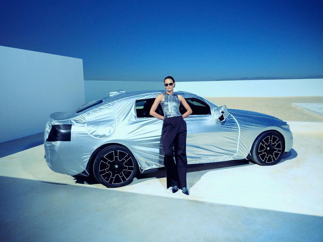 Fashion model poses outdoors next to a covered car under bright sky, wearing sunglasses and black wide-leg pants and a silver halter top.