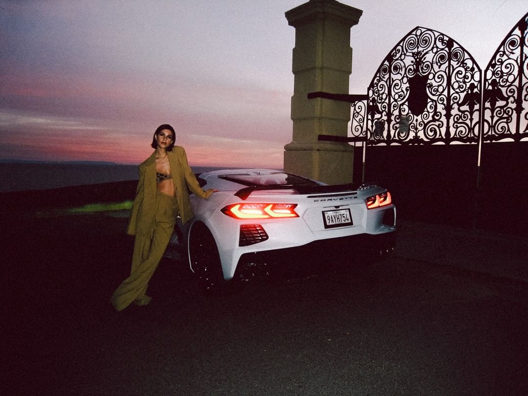 A woman in a tan suit poses with a white Corvette in front of an ornate gate against a dusky sky at twilight.