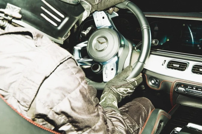 Person in driving suit grips steering wheel inside car with helmet, gloves, racing apparel; Mercedes logo visible; interior shot.