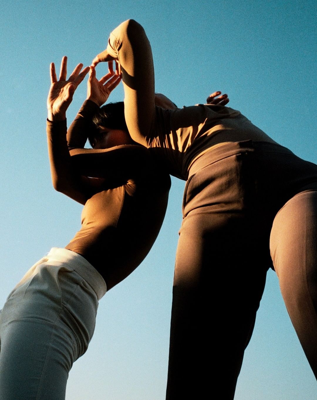 Two adults in modern, neutral outfits pose against a bright blue sky, creating dynamic silhouettes with their arms overhead.