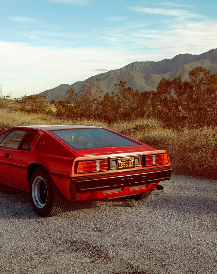 Red Lotus Esprit parked on a gravel road, mountains in the background under a partly cloudy sky, side rear angle view.