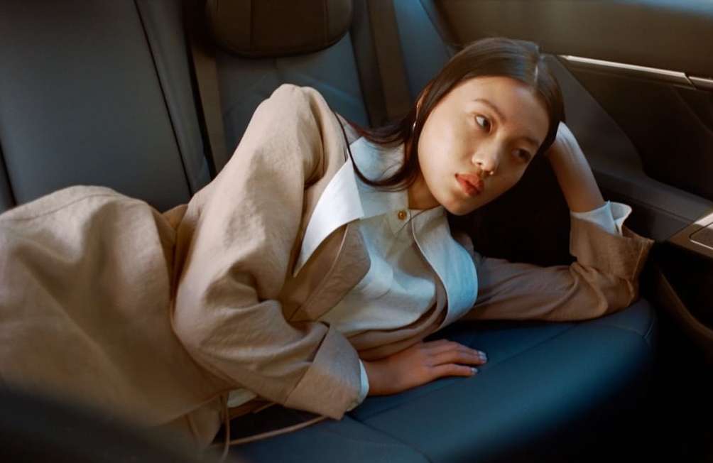 An Asian adult female reclines on the backseat of a car, wearing a tan blazer and white blouse, looking sideways with a neutral expression.