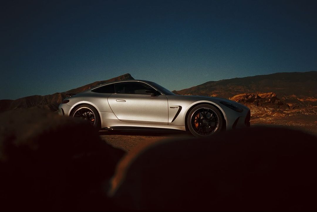 Silver Mercedes-AMG GT Coupe parked in a desert landscape under a clear blue sky, mountains in the background, car is the focus.