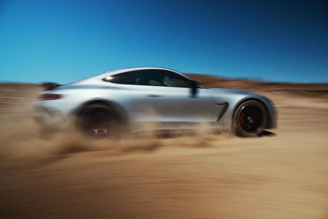 Silver sports car speeds across a tan desert landscape under a bright blue sky, kicking up sand in a motion-blurred shot.