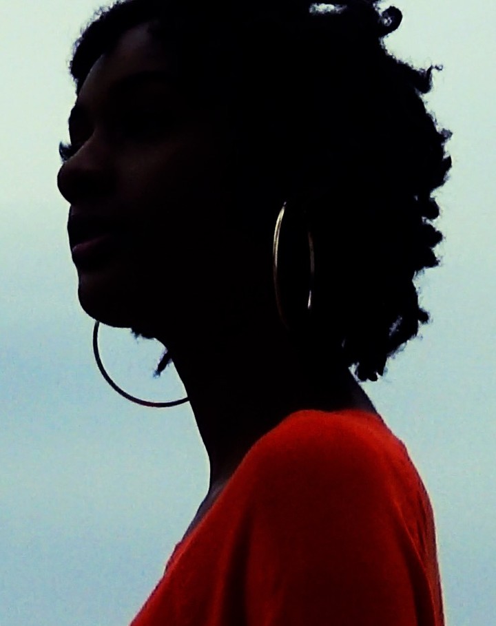 Close-up portrait of a Black woman in silhouette wearing large hoop earrings and a red top against a bright, overcast sky.