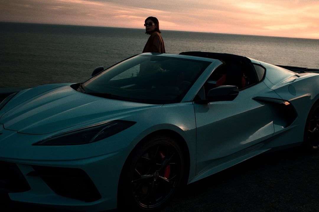 Convertible Corvette parked near the ocean at sunset with an adult female standing near the car looking toward the ocean view.