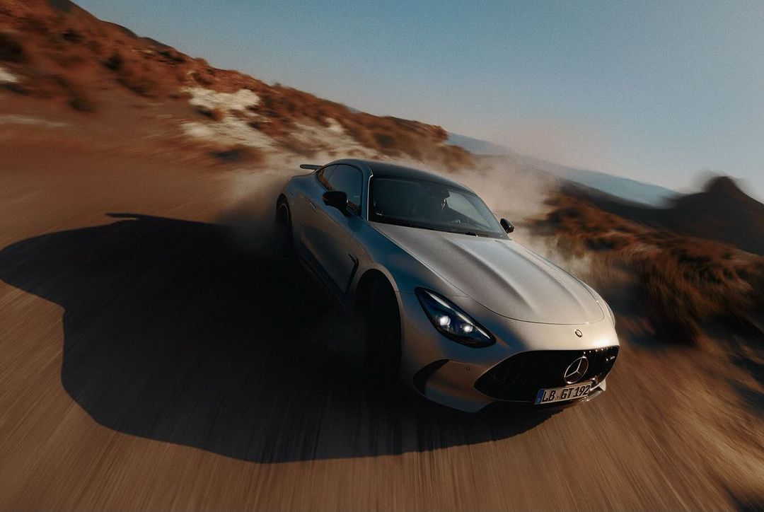 Silver Mercedes-AMG GT driving on a brown dirt road in a desert landscape, with a clear blue sky in the background during daylight.