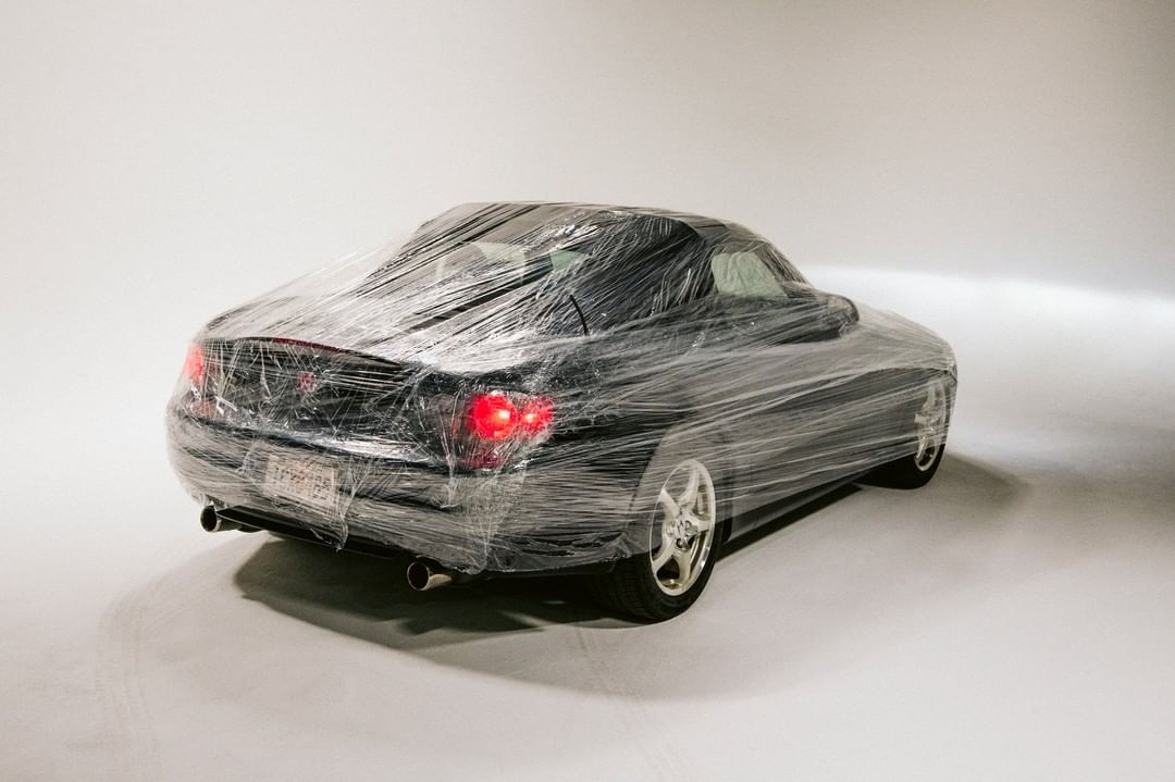 Black sports car is entirely encased in clear plastic wrap in a studio setting, lights illuminate the reflective surface. 
