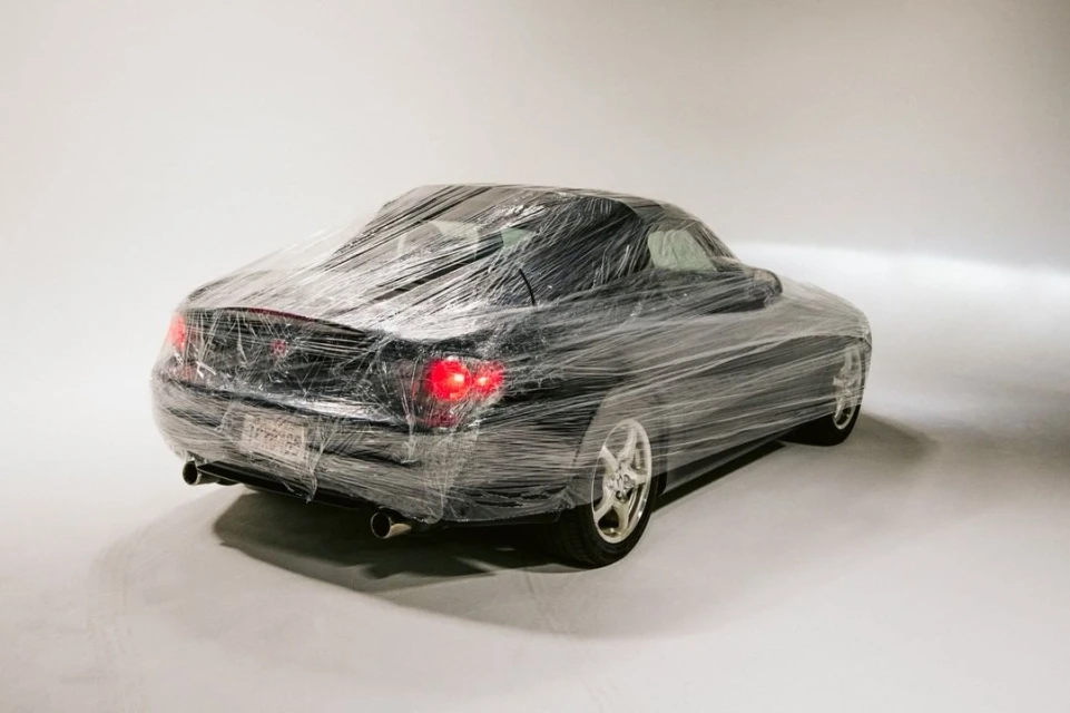 Black sports car is entirely encased in clear plastic wrap in a studio setting, lights illuminate the reflective surface. 