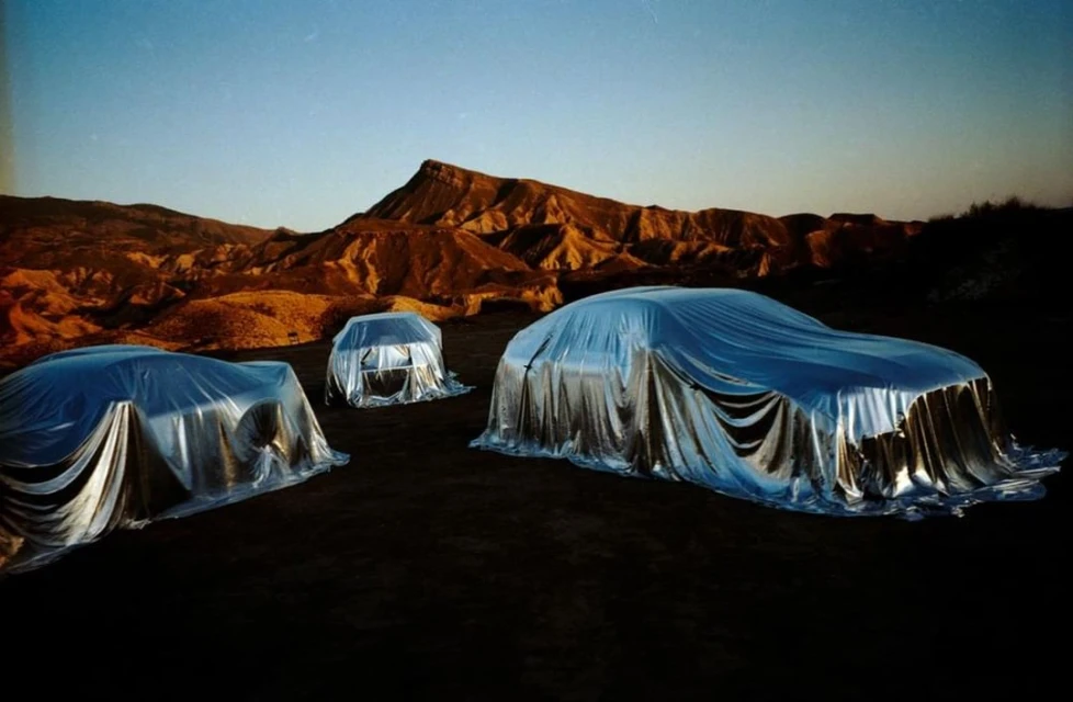 Three cars under silver wraps sit in a dark desert landscape, photographed at sunset against a backdrop of mesas and buttes.