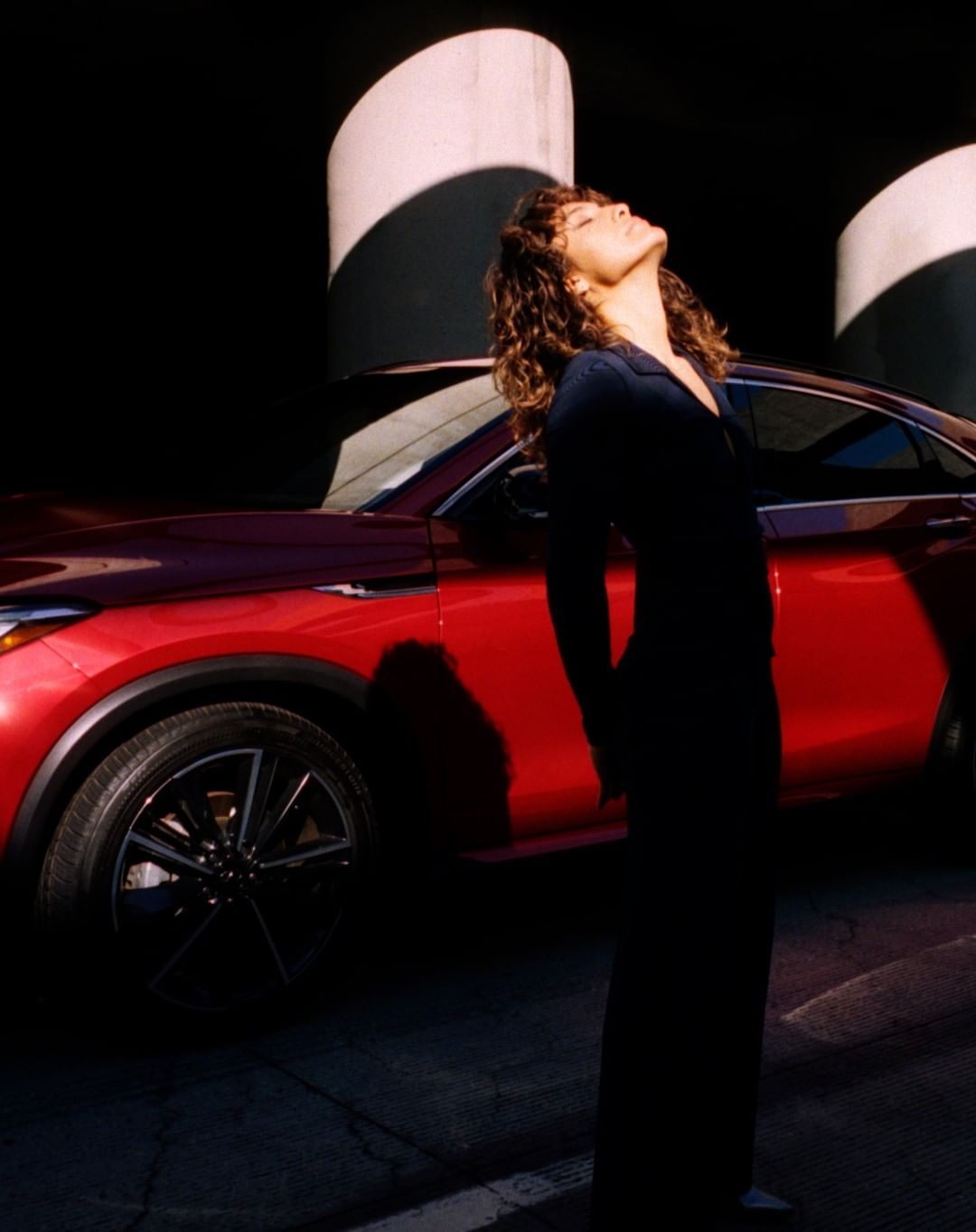 A woman with curly hair stands by a red SUV in an urban setting with pillars, basking in sunlight, creating a fashion-forward automotive image.