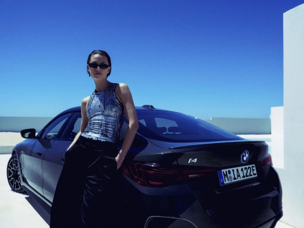 Brunette female model leans against a dark blue BMW i4 in a sunlit, minimalist outdoor setting with a clear blue sky overhead.