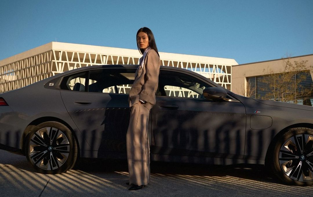 Woman in a suit poses next to a gray BMW 5 Series sedan on an outdoor city street with buildings under a blue sky at daytime.