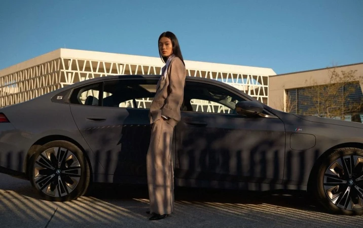Woman in a suit poses next to a gray BMW 5 Series sedan on an outdoor city street with buildings under a blue sky at daytime.