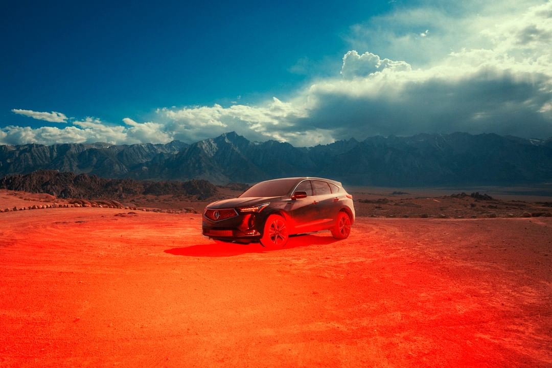 Black Acura RDX SUV on a reddish-orange desert landscape with mountains in the distance and a blue sky with clouds above. Automotive.