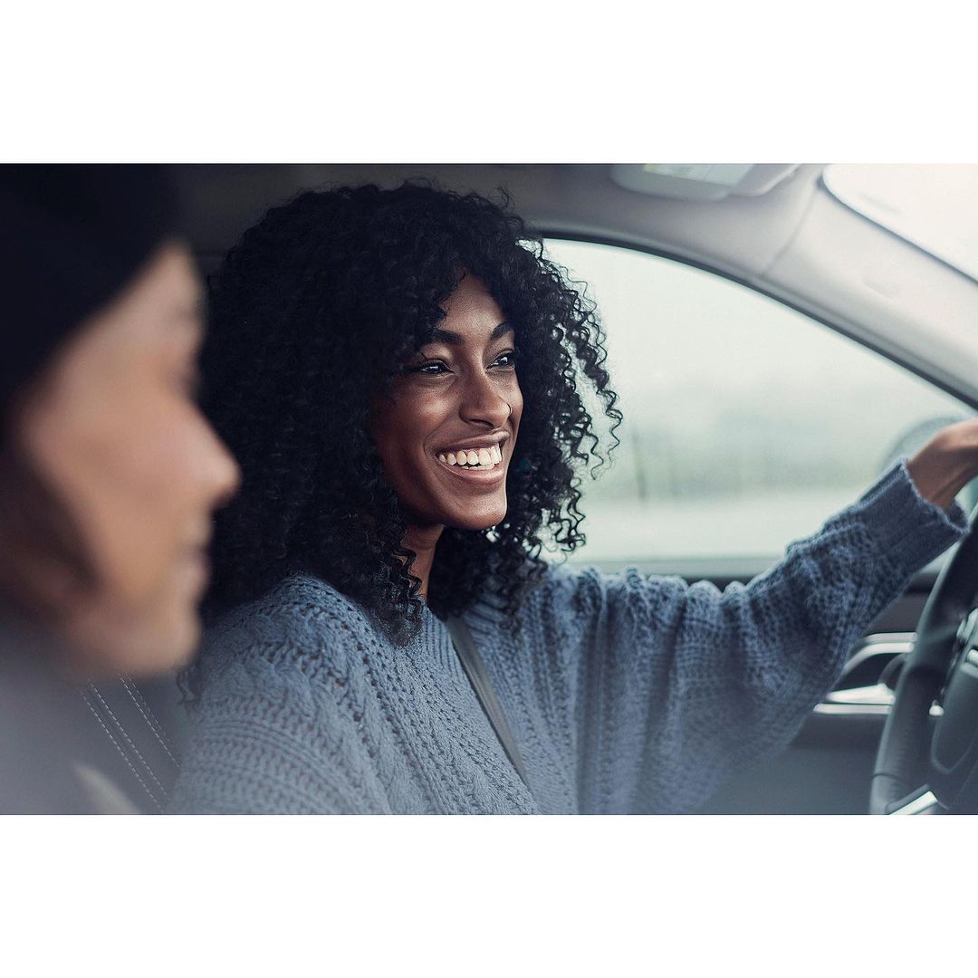 Two women inside a car, one driving and smiling with curly black hair, the other partially visible in the passenger seat.