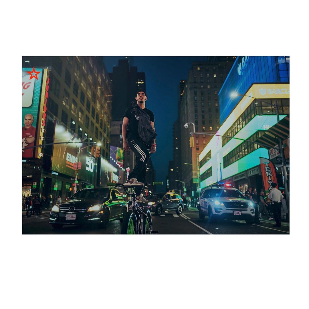 Man stands on a bicycle in Times Square at night with traffic, billboards, and buildings visible under a dark blue sky in an urban setting.