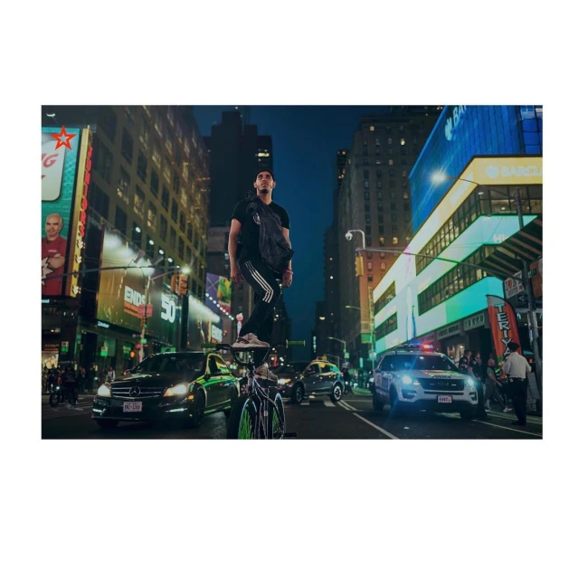 Man stands on a bicycle in Times Square at night with traffic, billboards, and buildings visible under a dark blue sky in an urban setting.