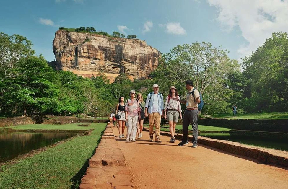 A group of tourists walking along a red dirt path in a tropical setting with a guide, with a large rock formation in the background.
