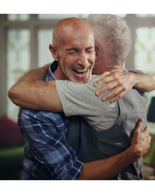 Two adult men embracing indoors. One man wears a blue plaid shirt and smiles with his eyes closed, while the other wears a gray shirt.