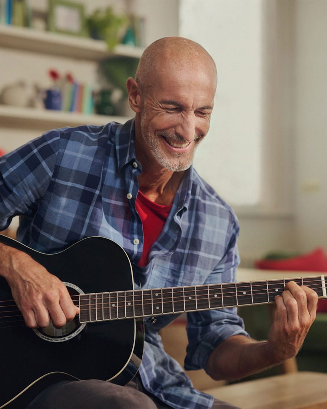 Adult man playing black acoustic guitar indoors, wearing blue plaid shirt and smiling, enjoying making music in casual setting.