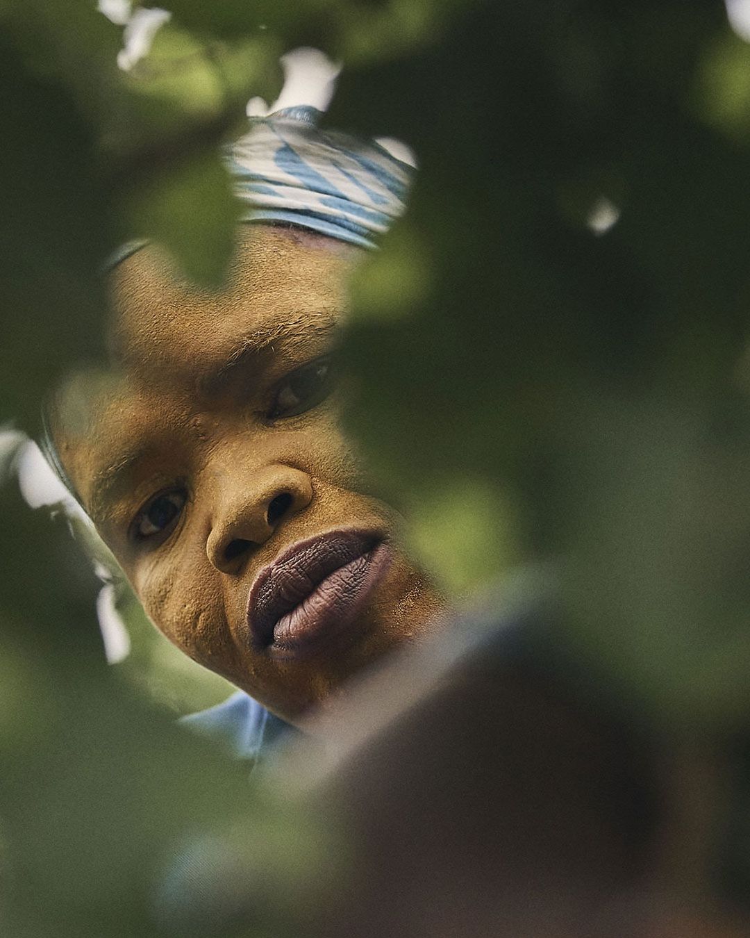 Close-up of an African woman with a headscarf, shot from a low angle, peering through lush green leaves in an outdoor setting.