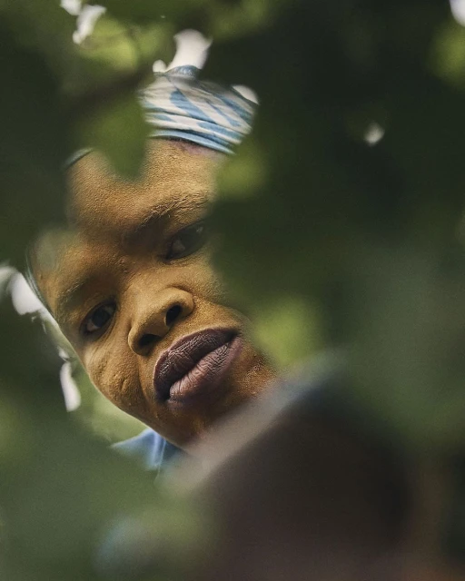 Close-up of an African woman with a headscarf, shot from a low angle, peering through lush green leaves in an outdoor setting.