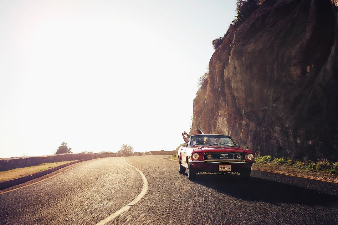 Classic red convertible car drives on a winding road near a rocky cliff on a sunny day, capturing a scenic automotive moment.