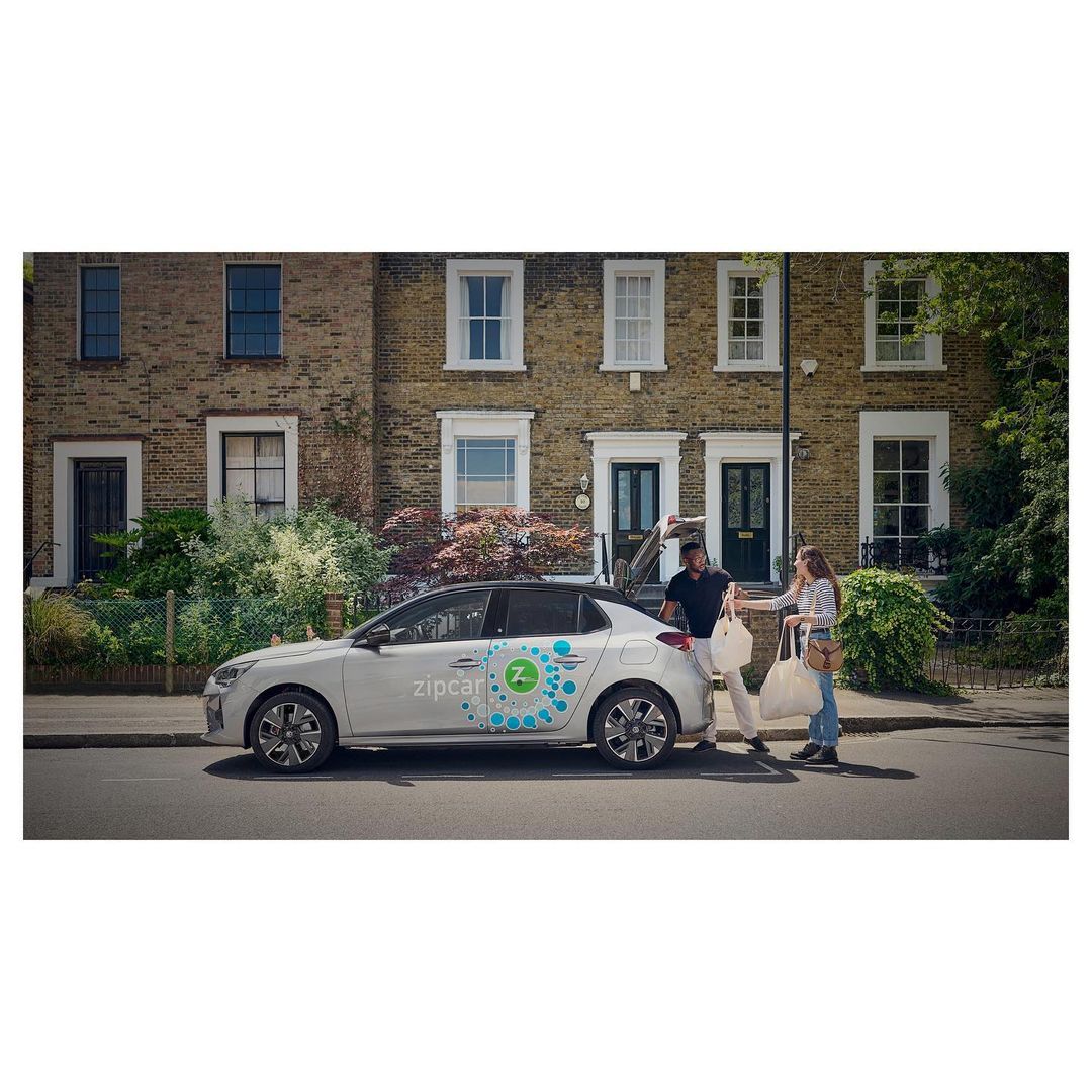Silver compact car with Zipcar logo parked on a city street with an adult male and female near the rear, carrying shopping bags.