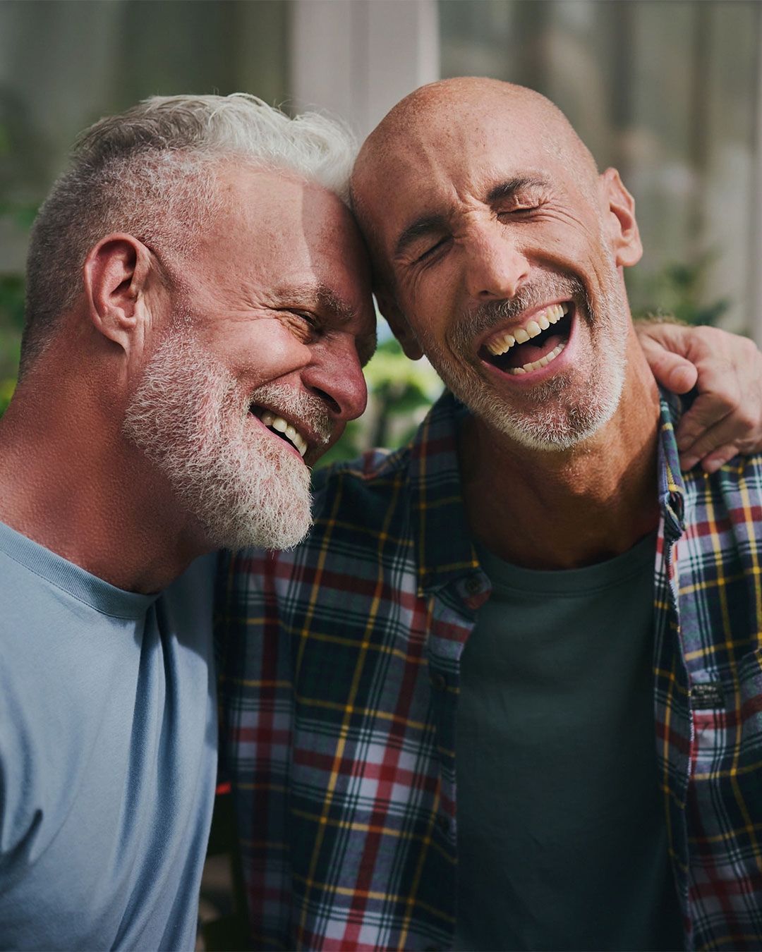 Two adult males smiling and laughing with their heads touching in an outdoor setting. The man on the right is wearing a plaid shirt.