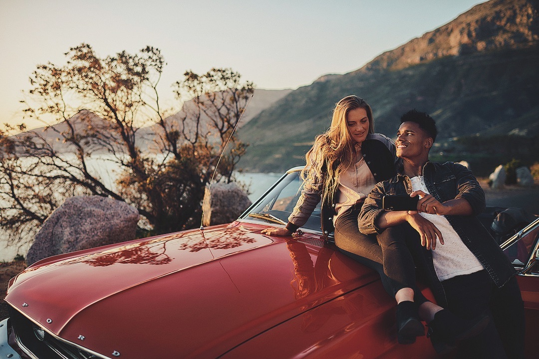 Couple poses on the hood of a red convertible car with mountains and a lake in the background, captured in warm, golden hour light.