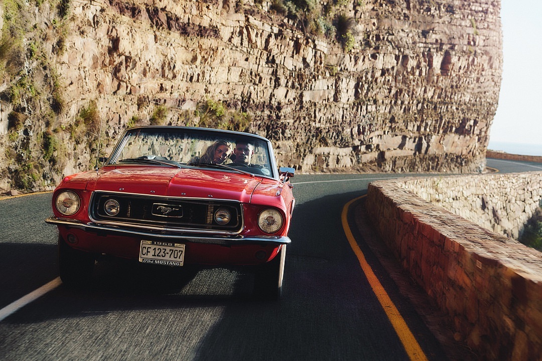 Red 1968 Ford Mustang convertible with two people driving on a coastal road beside a large rock face on a sunny day.