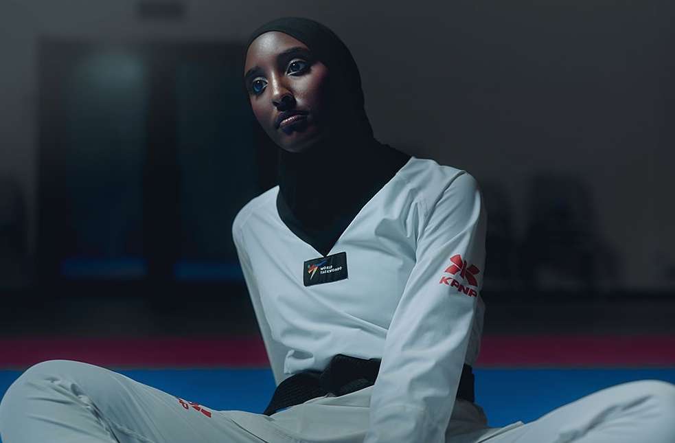 A portrait shows a woman in a white taekwondo uniform and black hijab sitting cross-legged on a blue mat, looking off to the side.