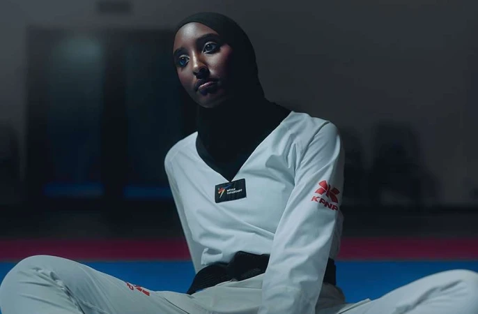 A portrait shows a woman in a white taekwondo uniform and black hijab sitting cross-legged on a blue mat, looking off to the side.