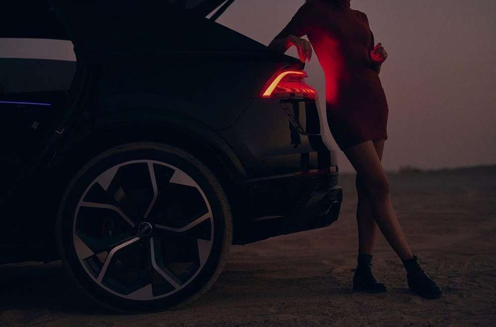 Woman in red dress leans on a black SUV with its trunk open, showcasing the car's taillight and large wheel against a dusky backdrop.
