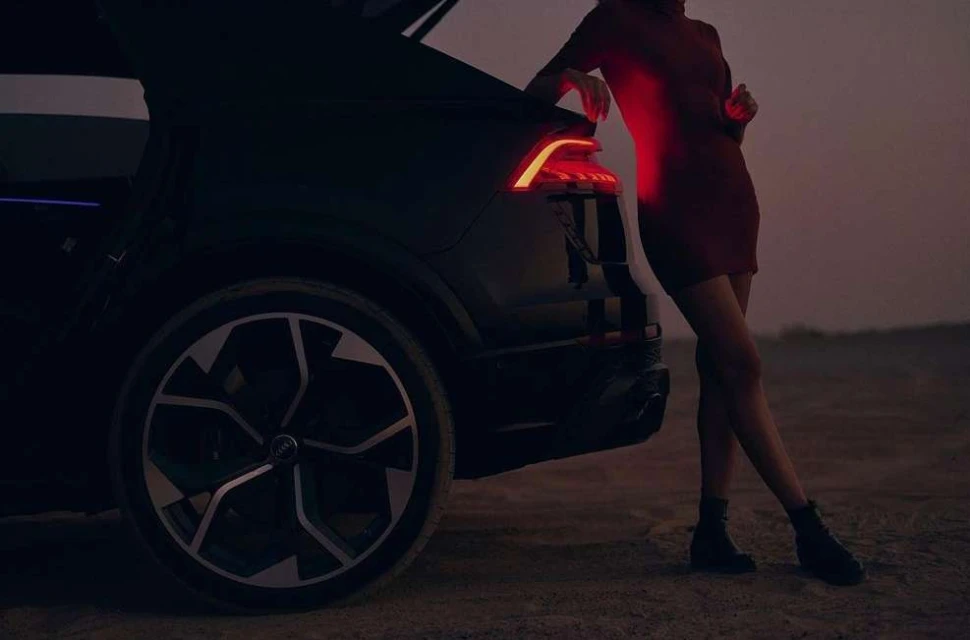 Woman in red dress leans on a black SUV with its trunk open, showcasing the car's taillight and large wheel against a dusky backdrop.