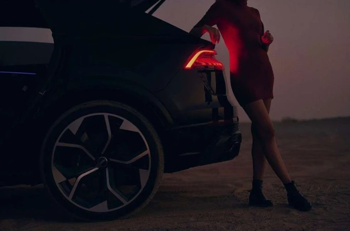 Woman in red dress leans on a black SUV with its trunk open, showcasing the car's taillight and large wheel against a dusky backdrop.