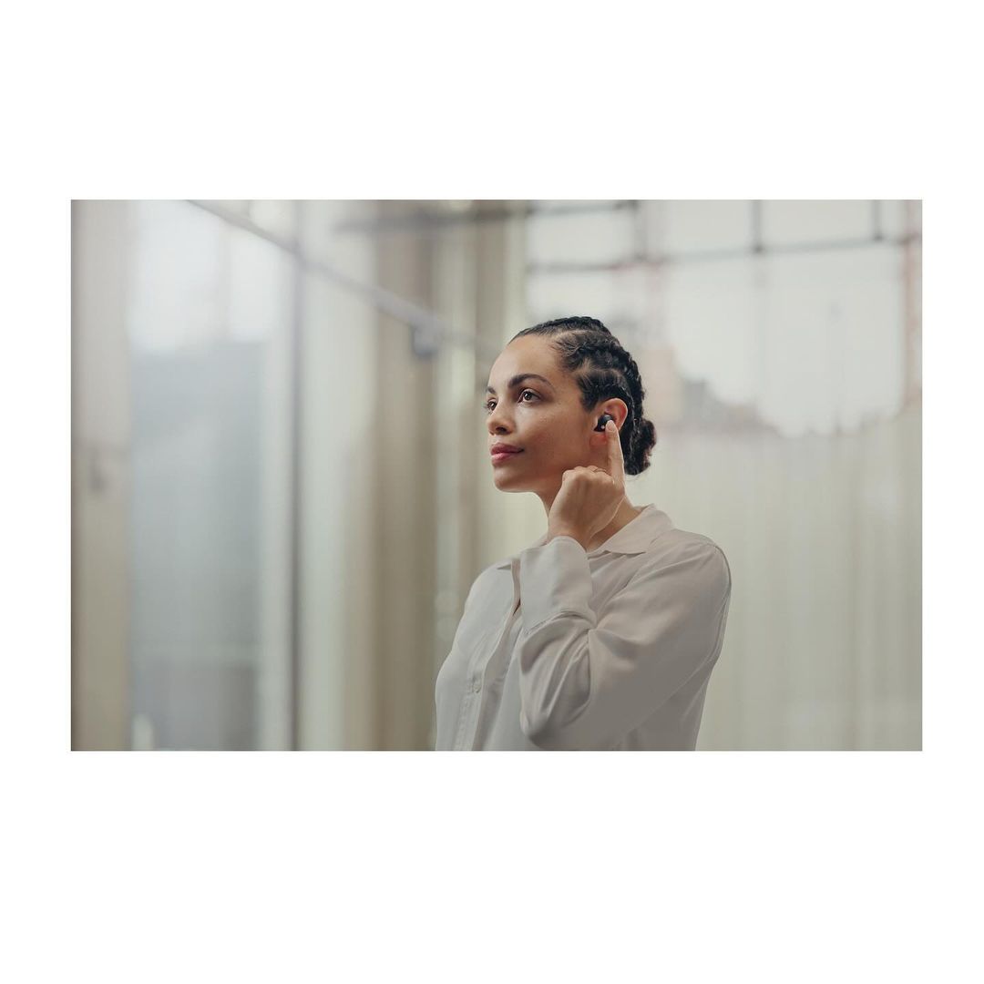 Woman with braided hair wearing wireless earbuds looking up and to the right in a brightly lit space with neutral walls.