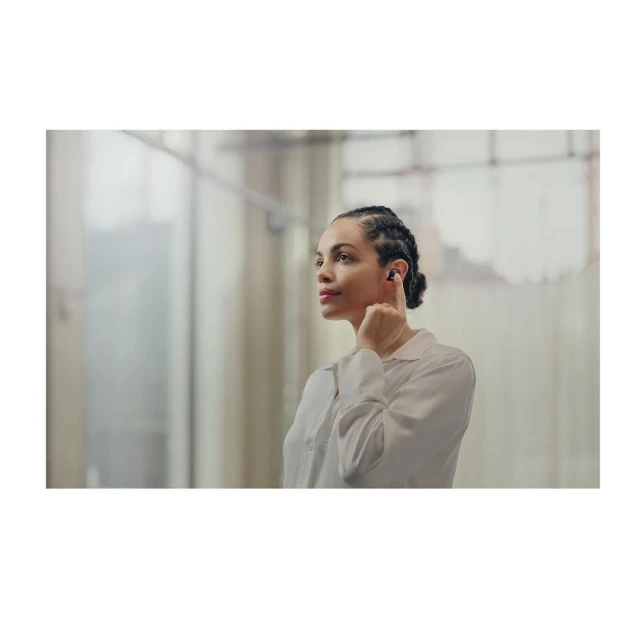 Woman with braided hair wearing wireless earbuds looking up and to the right in a brightly lit space with neutral walls.